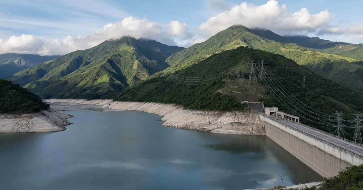 Embalse hidroeléctrico en los Andes colombianos con una franja clara de orillas expuestas, el coronamiento de la presa y líneas eléctricas sobre montañas verdes