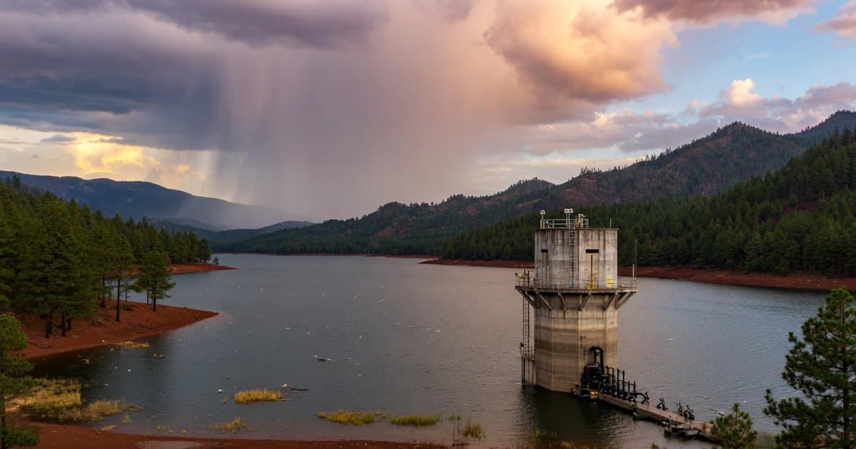 Gran embalse rodeado de montañas con bosques de pino en el centro de México, con el agua alta sobre la orilla y una torre de toma bajo nubes de tarde